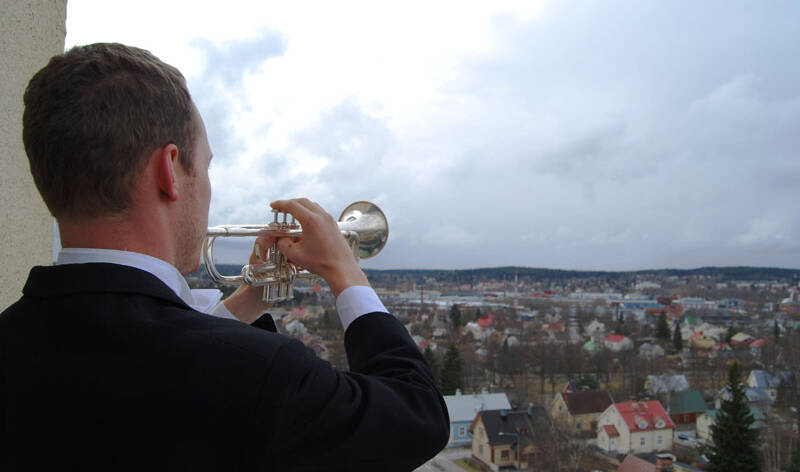 Mies seisoo selin ja soittaa trumpettia, joka on sivusuunnassa oikella. Taustalla kaupunkimaisesma, jossa on lähinnä rintamamiestaloja.