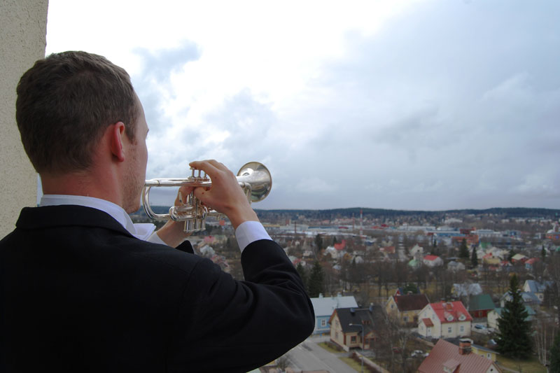 Mies seisoo selin ja soittaa trumpettia, joka on sivusuunnassa oikella. Taustalla kaupunkimaisesma, jossa on lähinnä rintamamiestaloja.