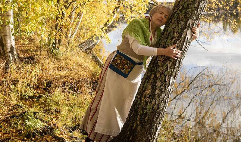 Nainen nojaa puuhun syksyisessä maisemassa järven rannalla.