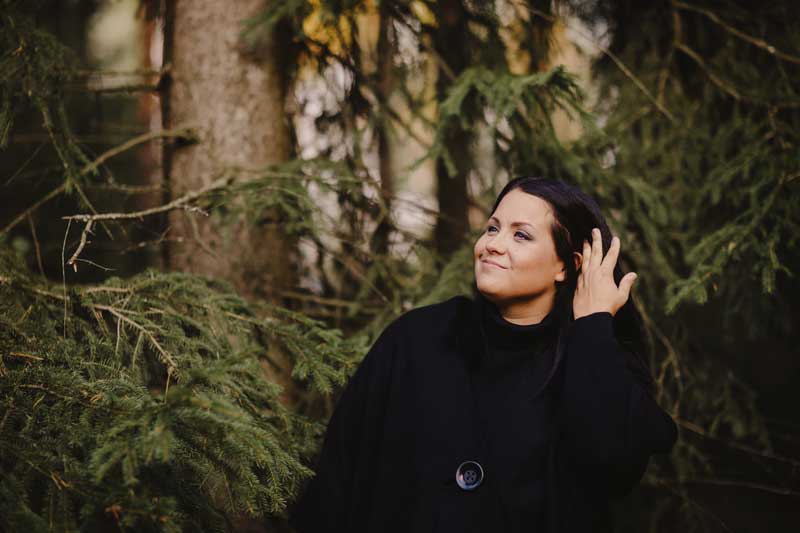 Henkilökuva Hanna Kivisalosta, joka seisoo vanhojen kuusien edessä.
