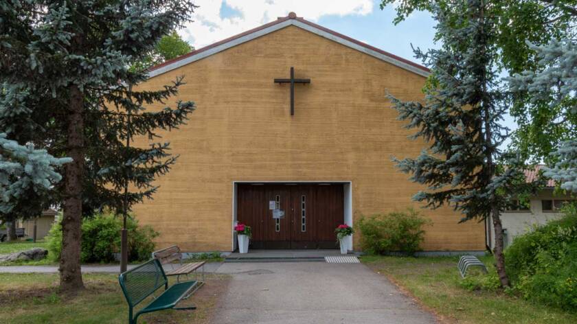 Above the dark doors of the light colored church building, there is a brown wooden cross.