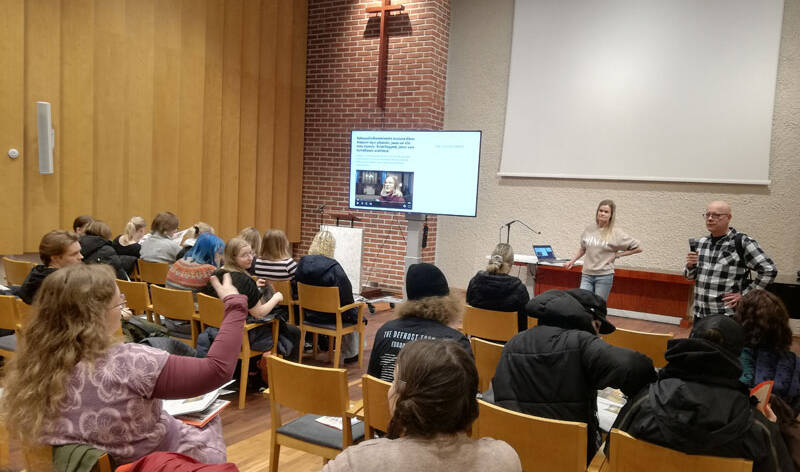 Joukko nuoria istuu salissa, ja heidän edessään seisoo nainen ja mies mikrofonien kanssa.