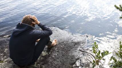 Huolestuneen näköinen henkilö istuu yksin kivellä, järven rannalla.