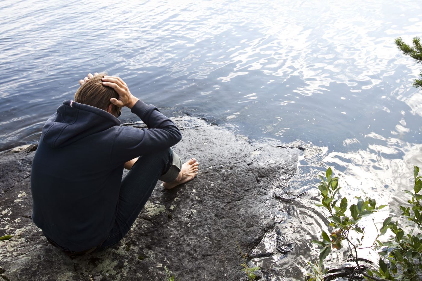 Huolestuneen näköinen henkilö istuu yksin kivellä, järven rannalla.