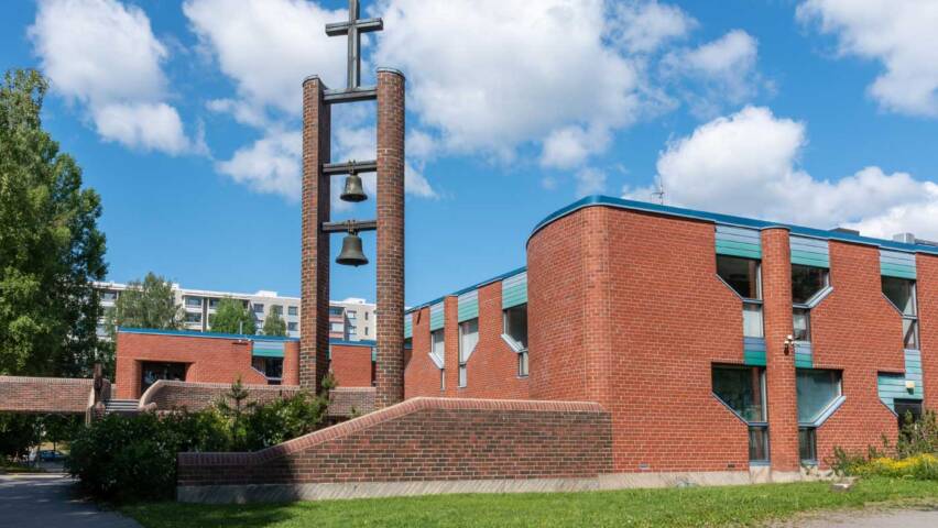 Behind the red brick bell tower is a low, rounded-cornered red brick church building shaded by green leafy trees.