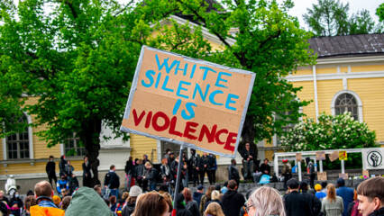Suuri väkijoukko ulkona Tampereen keskustorilla. Väkijoukossa yhdellä on suuri kyltti, jossa lukee englanniksi: White silence is violence.