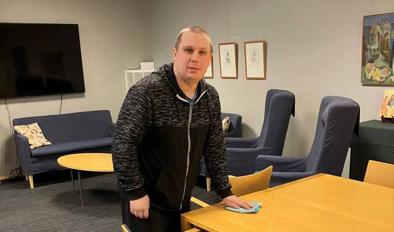 Nuorimies seisoo pöydän ääressä kokoustilassa ja pyyhkii pöytää.