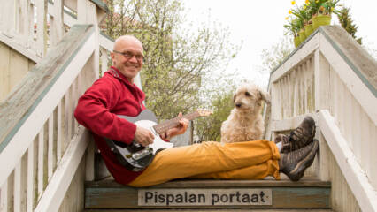 Mies istuu puisillaportailla, joissa lukee Pispalan portaat, kitara sylissä ja pieni koitra katselee vieressä.