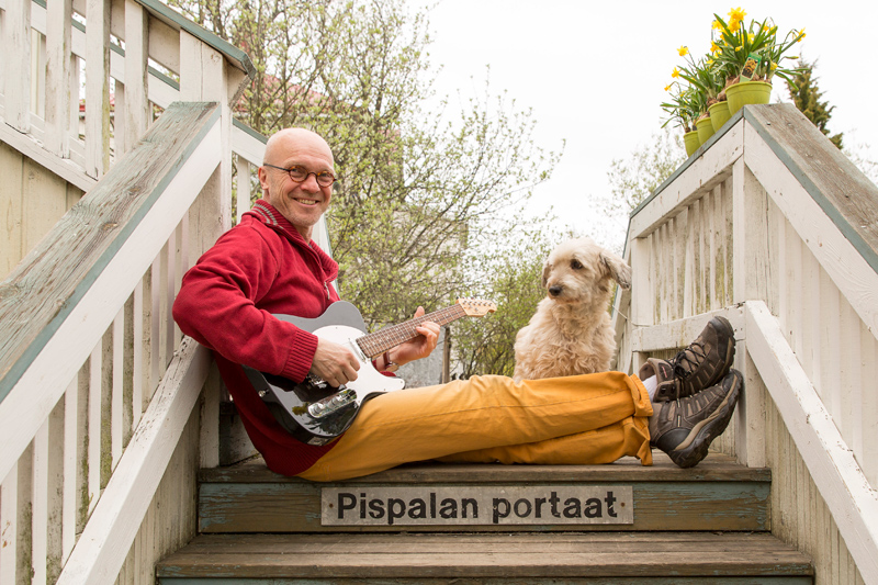 Mies istuu puisillaportailla, joissa lukee Pispalan portaat, kitara sylissä ja pieni koitra katselee vieressä.