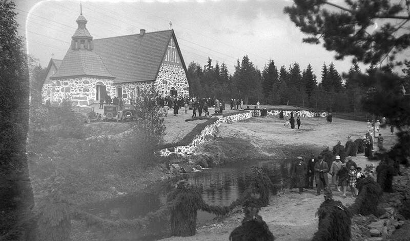 Vanha ja osin vaurioitunut kuva kirkosta, jonka edustalla on ihmisiä ja vanhoja autoja. Edessä virtaa joki, jonka vieressä on heinäseipäistä ja niiden päällä olevista köynnöksistä muodostettu kuja, jota pitkin kulkee ihmisiä. Seipäillä on heinää.