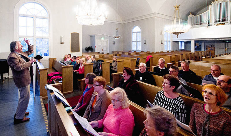 Kuoronjohtaja Vesa Haapaniemi on kuoron edessä ja kuorolaiset istuvat Vanhan kirkon penkeissä.