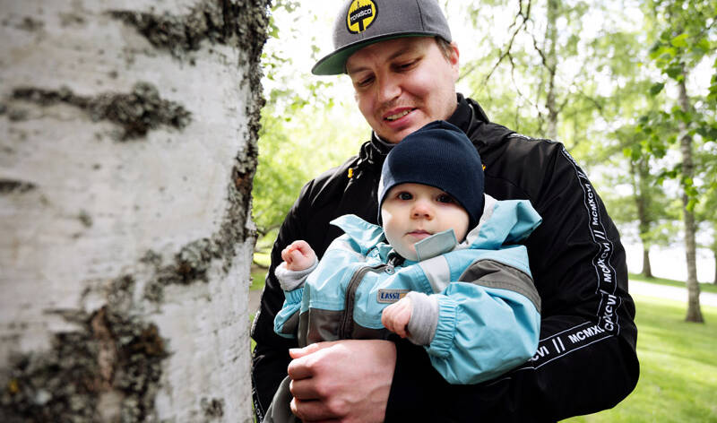 Isä vauva sylissään ulkona koivun vieressä.