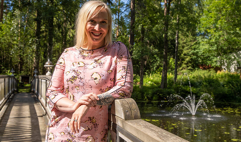 Aurinkoinen kesäpäivä. Hymyilevä Jutta Larm nojaa puusillan kaiteseen. Taustalla pieni suihkulähde suihkuaa joessa.