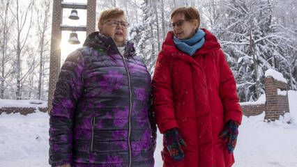 Kaksi naista seisovat talvisessa säässä Hervannan kirkon ovilla. Takana näky kellotorni, joiden takaa aurinko paistaa.