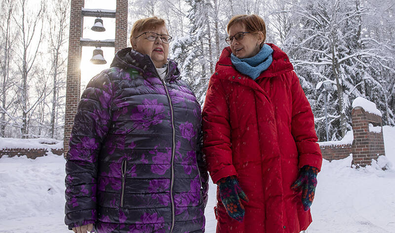 Kaksi naista seisovat talvisessa säässä Hervannan kirkon ovilla. Takana näky kellotorni, joiden takaa aurinko paistaa.