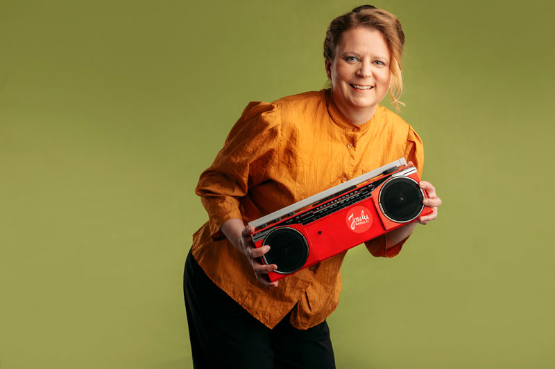 Iloisesti hymyilevä henkilö pitää käsissään punaista radiomankkaa, jossa lukee jouluradio.