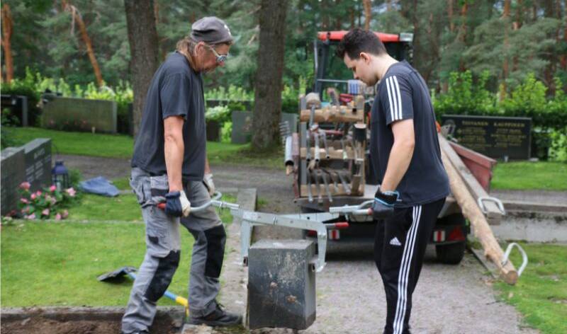 Kaksi miestä kantaa haudan reunuskiveä.
