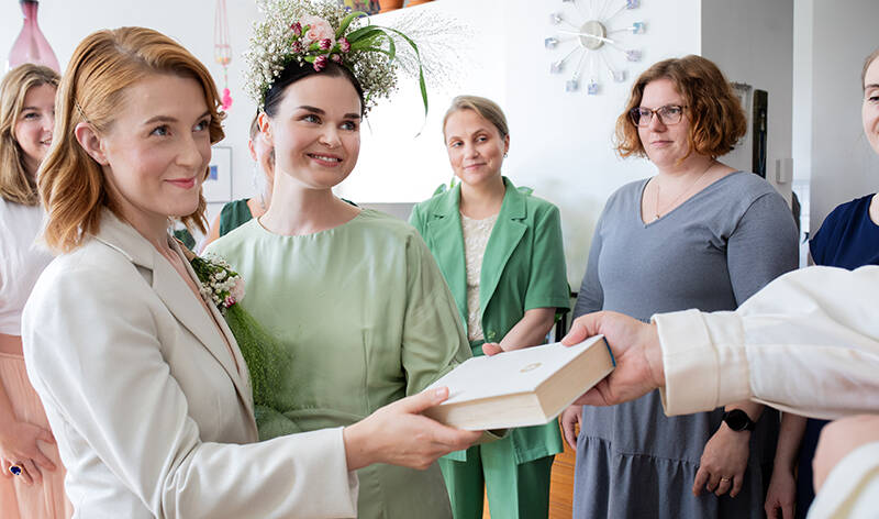 Vierekkäin on kaksi naista, joille käsi ojentaa valkoista kirjaa. Toisella naisista on kukkapäähine ja toisella kukkia rintapielessä. Taustalla seisoo viisi naista.