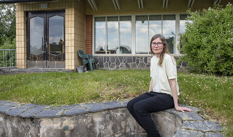 Hymyilevä henkilö istuu ulkona kivisellä kaiteella, ja taustalla on rakennus.