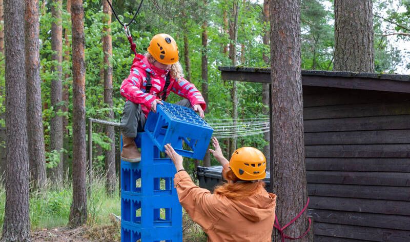Lapsi on kiivennyt sinisten juomakorien päälle, päässääm oranssi turvakyparä.Aikuinen onselin ja pitää häntä käsistä Lielahden kirkon kesäleiripäivässä.