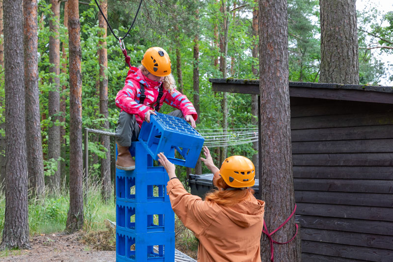 Lapsi on kiivennyt sinisten juomakorien päälle, päässääm oranssi turvakyparä.Aikuinen onselin ja pitää häntä käsistä Lielahden kirkon kesäleiripäivässä.