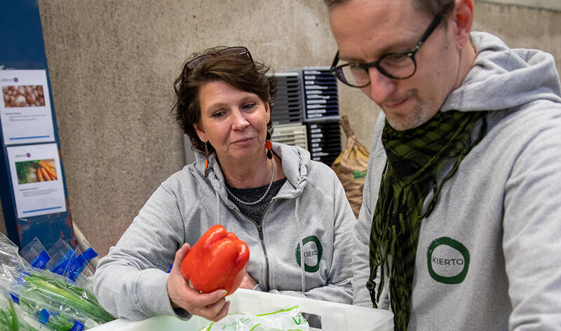 Nainen ja mies ruokalaatikoiden edessä katselemassa vihanneksia. Naisella kädessä punainen paprika.