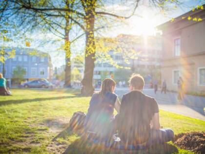 Nuoripari istuu nurmikkolla kevään vihreiden puiden alla ,ja aurinko on laskemassa heidän edessä olevien talojen taakse.
