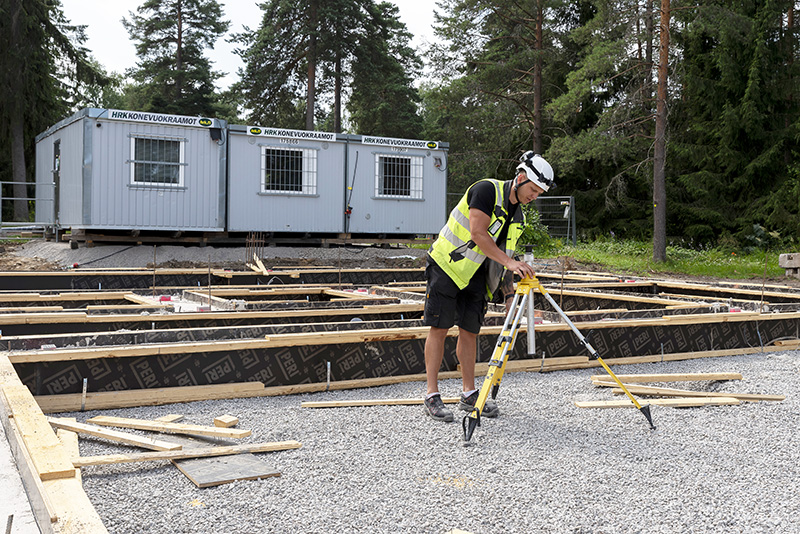 Henkilö asettelee kolmijalkaa rakennuksen pohjarakenteen edessä, ja takana on parakkimainen rakennus.