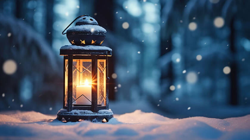 Tummassa metallisessa kynttilälyhdyssä, joka on lumisessa maassa palaa valkoinen kynttilä, taustana tumma luminen taivas.