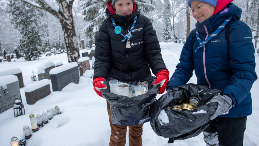 Kaksi henkilöä keräävät kynttilöitä lumisella hautausmaalla, ja he pitävät sylisään mustia jätesäkkejä, joissa on useita palaneita kynttilöitä.