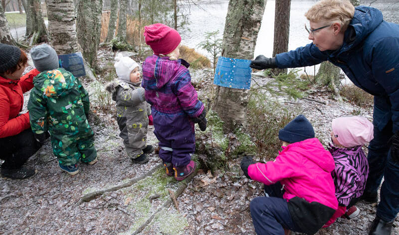 Ryhmä pieniä lapsia ja kaksi aikuista tutkii tehtävälappua marraskuisessa säässä metsässä järven rannalla.