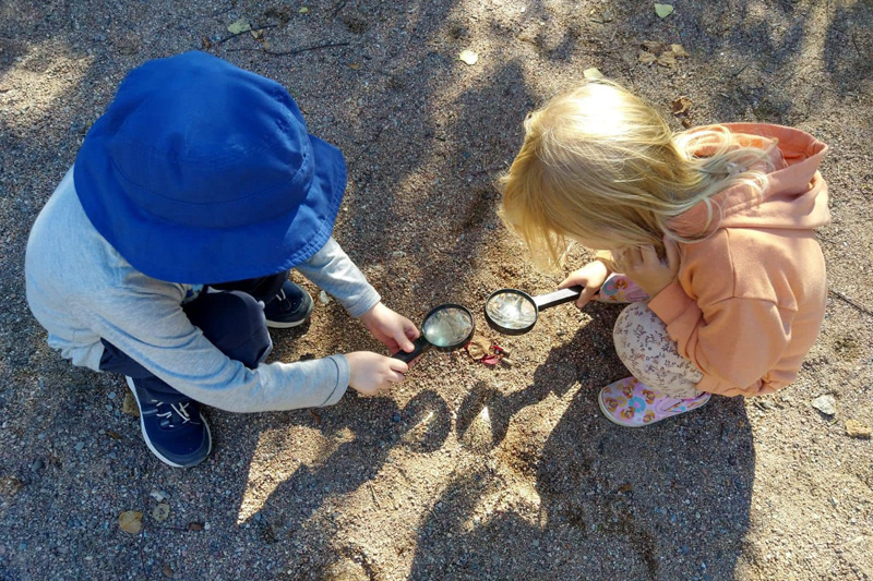 Kaksi lasta pihalla kyykistyneenä tutkimaan pihaa suurennuslasien kanssa.