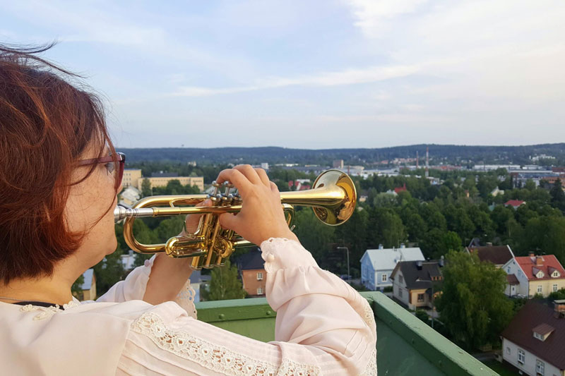 Nainen seisoo selin ja soittaa trumpettia, joka on sivusuunnassa oikella. Taustalla kaupunkimaisesma, jossa on lähinnä rintamamiestaloja.