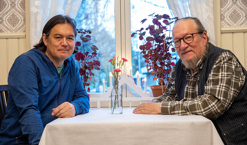 Kaksi miestä istuu ikkunan edessä olevan kotoisan oloisen pöydän ääressä. Pöydällä on kukkia lasipullossa, ja ikkunan edessä kynttelikkö ja kasveja.