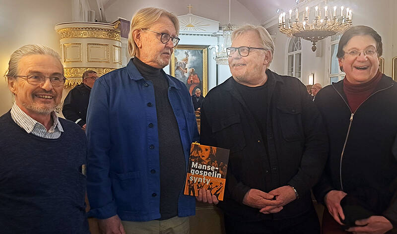 Neljä miestä seisoo vierekkäin, ja yksi heistä pitää kirjaa esillä. Oikeanpuoleinen heistä nauraa leveästi.