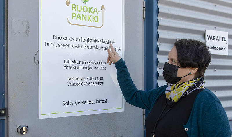 Nainen seisoo ruokapankin ovella ja osoittaa info-tekstejä.