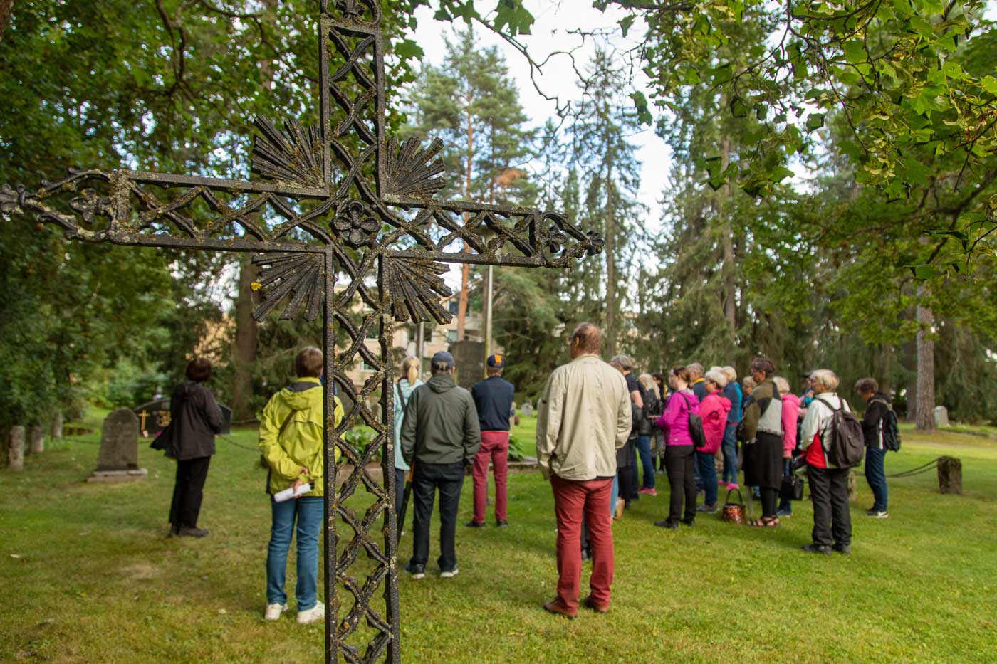 Kesäisiin vaatteisiin pukeutuneet ihmiset seisovat ison rautaristihautamuistomerkin takana.