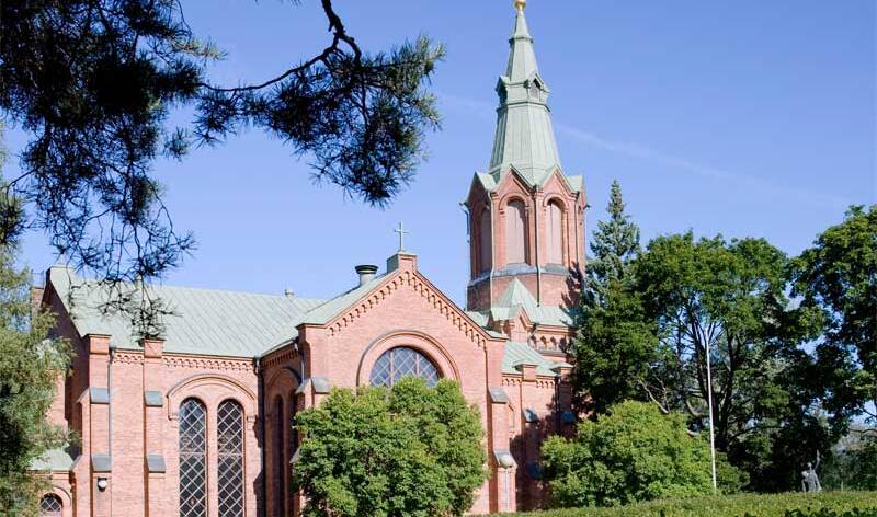 Punatiilistä kirkkorakennusta ympäröivät korkeat lehtipuut ja kasvava nurmialue.
