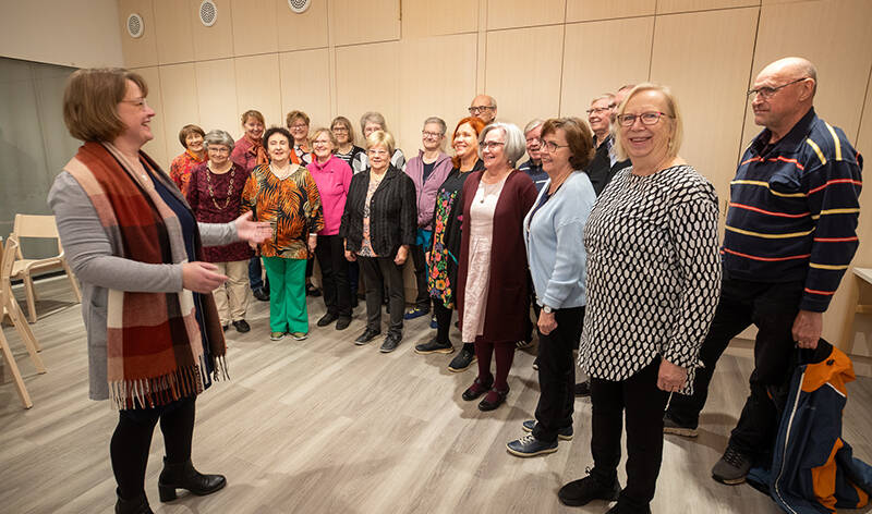 Joukko iloisia ihmisiä seisoo puisessa salissa, jonka seinällä on risti. Vasemmalla puolella erillään muista seisova nainen heiluttaa käsiään.