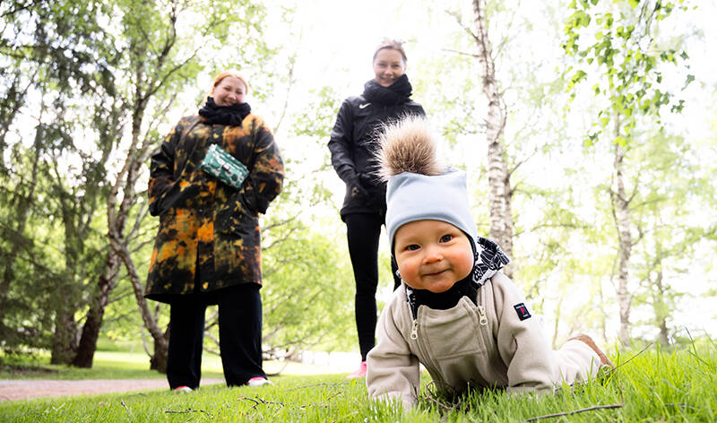 Vauva ryömii vihreällä nurmella kohti. Aikuiset katselevat taustalla.