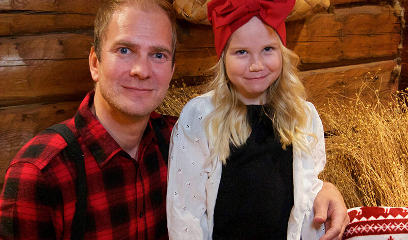 Ruutupaitainen mies ja rusettilakkinen tyttö istuvat vierekkäin hirsiseinän edessä sisällä. Seinällä on tuohikontti ja vieressä kuivattuja kasveja.