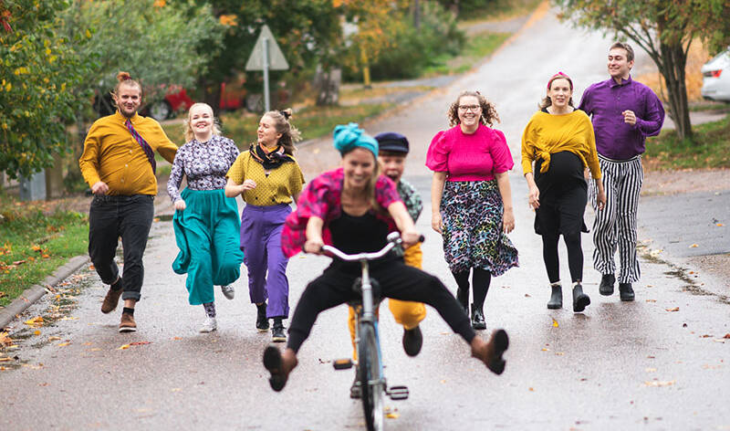Kahdeksan ihmistä värikkäissä ja hauskoissa vaatteissa juoksee tiellä riemastuneina.