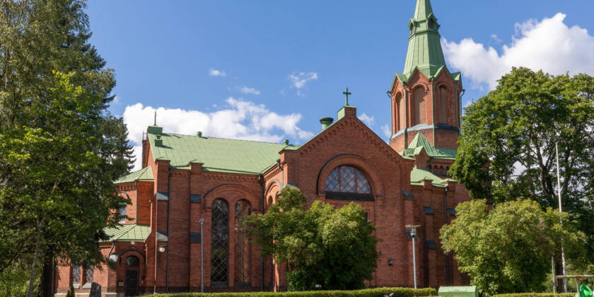 Yleiskuva Messukylän kirkosta, joka on uusgoottista tyyliä edestava punatiilinen kirkko ja kirkko on  vihreiden lehtipuiden ympäröimänä ja etualalla hautausmaan hautakiviä.