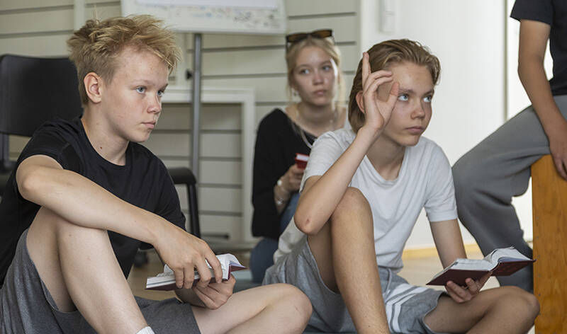 Kolme nuorta istuu kirjat käsissään, ja yksi heistä viittaa. Kuvan reunassa näkyy neljännen henkilön jalka.