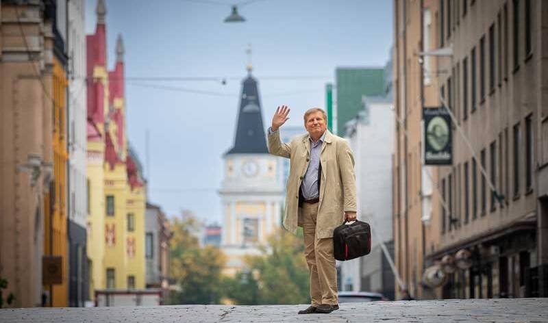 Mies vaaleassa popiilinitakissa seisoo keskellä katua, ja hänellä on toisessa kädessä salku ja toisella hän heiluttaa, taustalla näkyy keltainen kirkko.