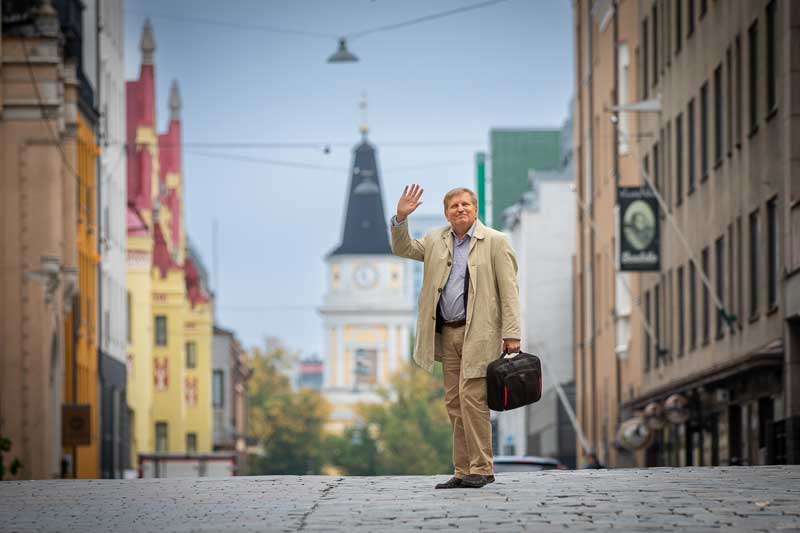 Mies vaaleassa popiilinitakissa seisoo keskellä katua, ja hänellä on toisessa kädessä salku ja toisella hän heiluttaa, taustalla näkyy keltainen kirkko.