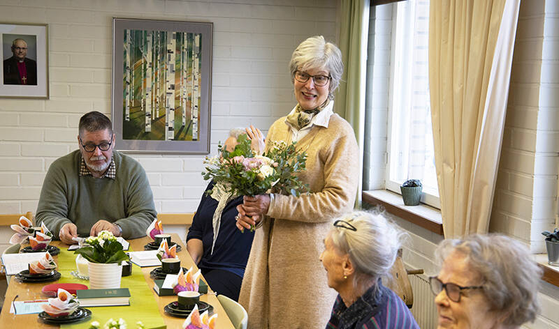 Kahvipöydän ääressä on ihmisiä, joista yksi seisoo kukkakimppu kädessään.