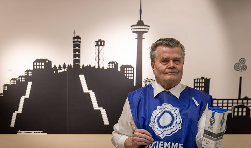 Mies lipas kädessä, sininen liivi päällä, taustalla seinällä siluetti Tampereesta.