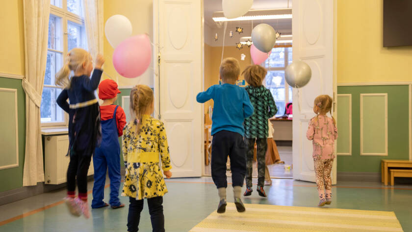 Joukko lapsia juoksee kohti salin avointa ovea ja hyppii ilmaan ilmpallot käsissään.