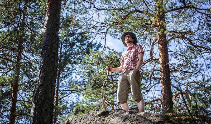 Mies seisoo kalliolla keppi kädessä ja männyt taustalla.